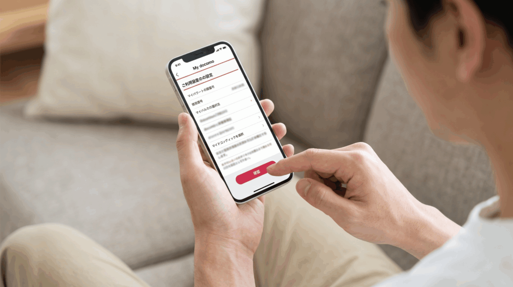 リビングのソファでリラックスしながら、スマホのMy docomoアプリ画面でプラン変更手続きの決定ボタンをタップする日本人男性。